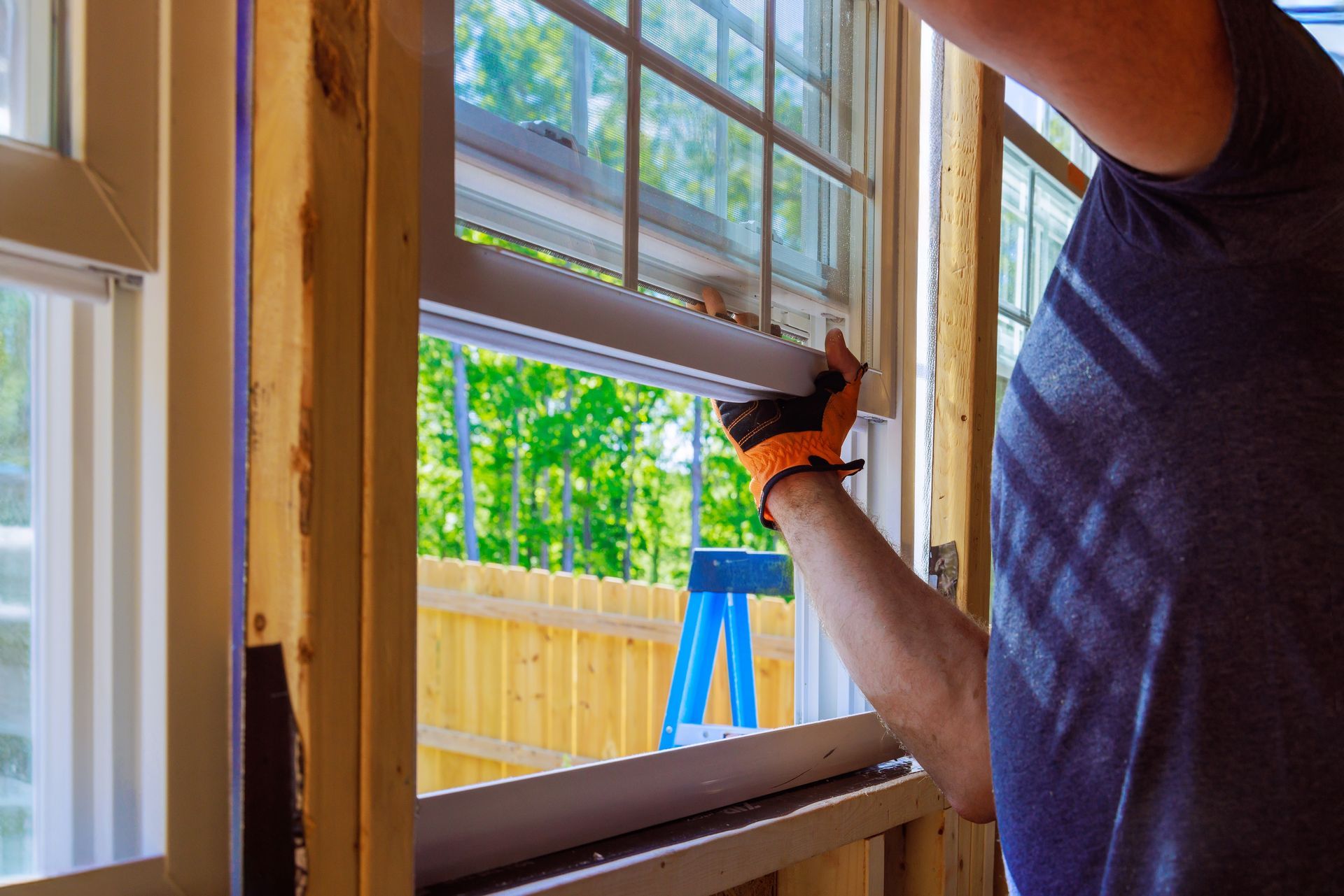 Person installing a white window, wearing gloves, in a wooden frame; background shows trees and a blue ladder.
