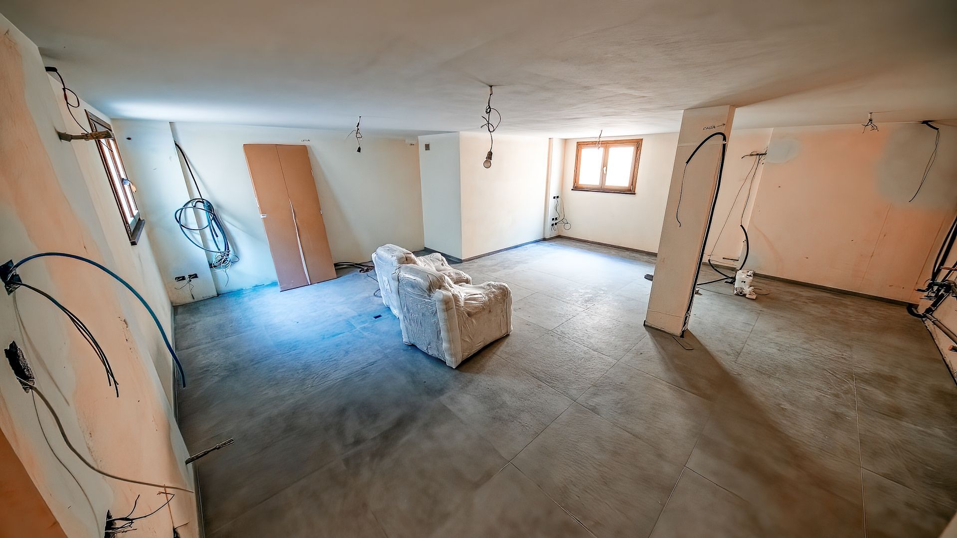 Interior of a room under construction; exposed wires on walls, unfinished walls, a wrapped sofa, concrete floor.