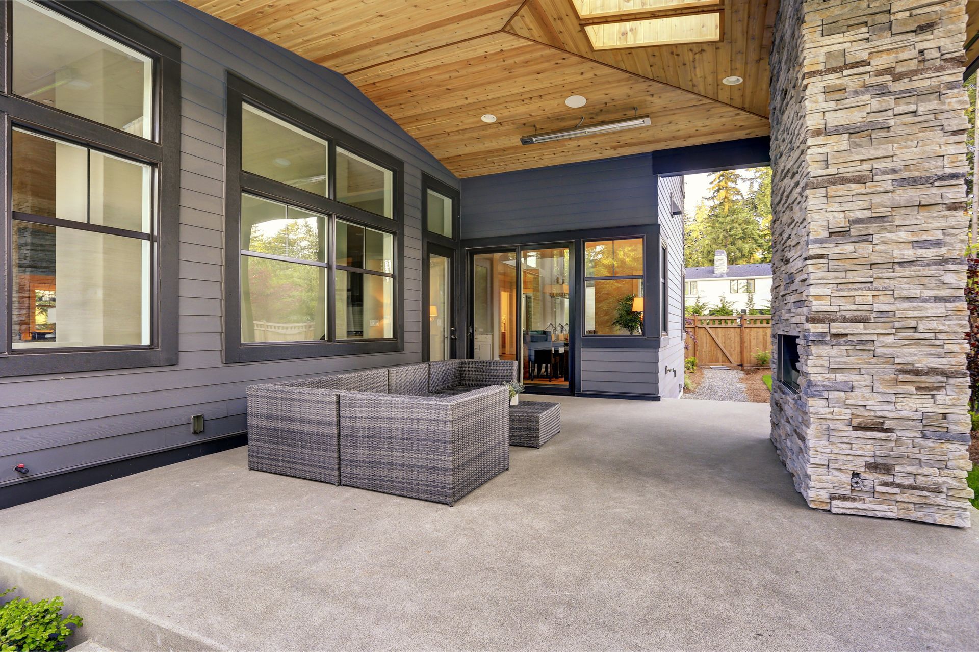Gray patio with wicker furniture, gray siding, stone fireplace, and wood ceiling.