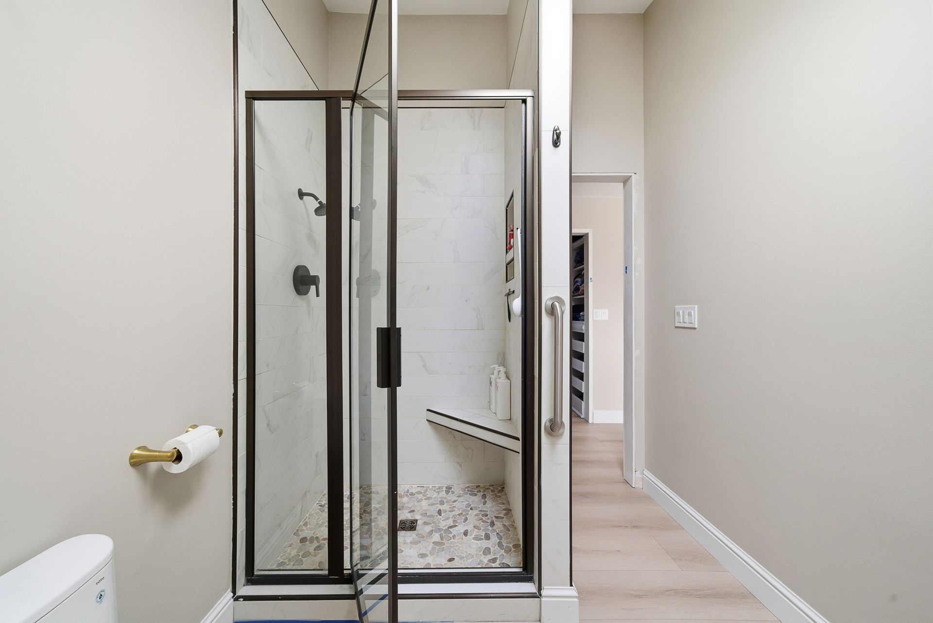 A walk-in shower with dark frames, a pebble floor, a built-in bench, and a grab bar in a neutral-toned bathroom.