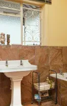 A bathroom with brown tile walls, a white pedestal sink, a bathtub, a grey storage cabinet, and a window with a metal grill.