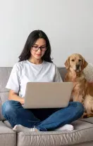 A person sits cross-legged on a sofa, working on a laptop with two golden retrievers nearby in a bright, modern room.