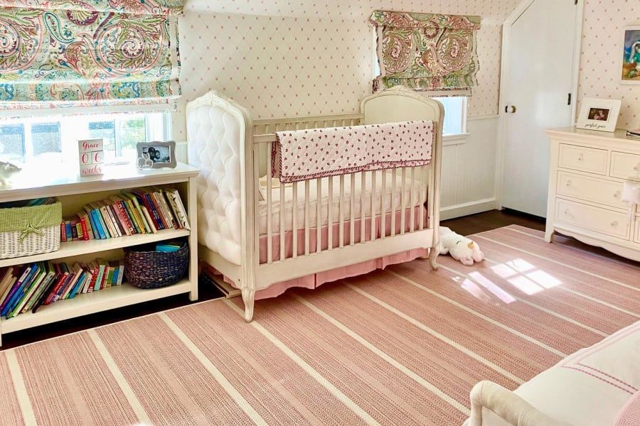A nursery with a white tufted crib, a bookshelf filled with books, a white dresser, and a pink-and-white striped rug.