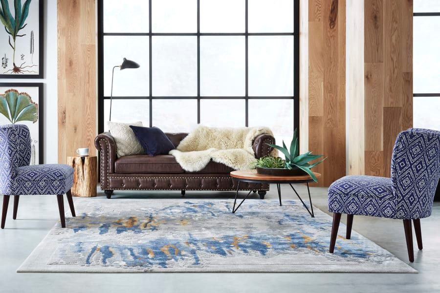 A living room featuring a brown leather sofa, two patterned purple chairs, and a blue and grey abstract rug.