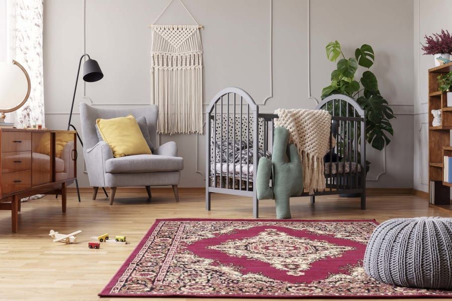 A nursery featuring a grey crib, a light grey armchair with a yellow pillow, a patterned burgundy rug, and a cactus pillow.