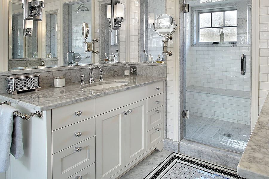 White bathroom vanity with marble countertop and sink, next to a glass-enclosed shower with tiled walls and flooring.