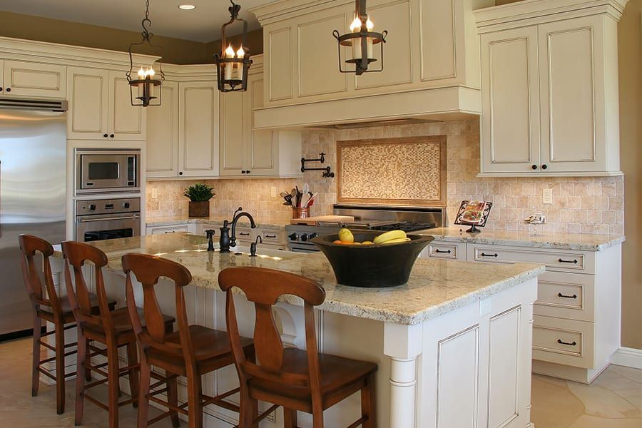 A kitchen with white cabinets, a large island with four wooden stools, granite countertops, and three hanging lanterns.