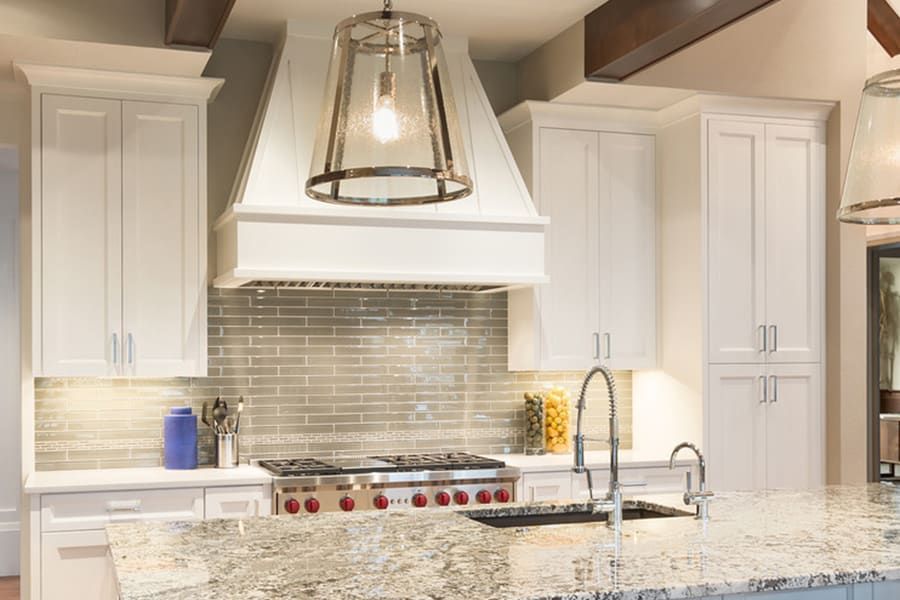 A modern kitchen with white shaker cabinets, a gray subway tile backsplash, a large vent hood, and granite countertops.