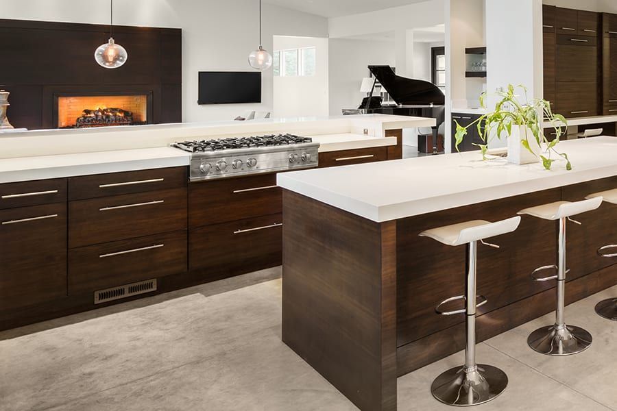Modern kitchen with dark wood cabinets, white countertops, stainless steel stove, and bar stools on a tiled floor.