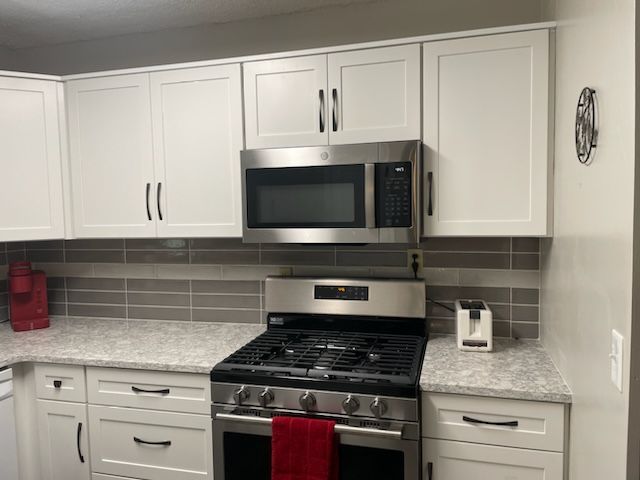 White kitchen cabinets with black handles, a stainless steel microwave and gas stove, and a grey tile backsplash.