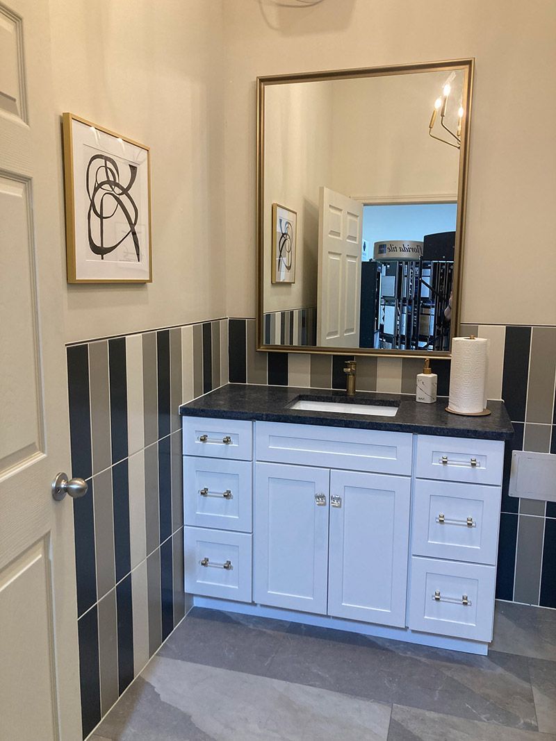 A white bathroom vanity with black countertop and striped tile backsplash, featuring a gold-framed mirror and wall art.