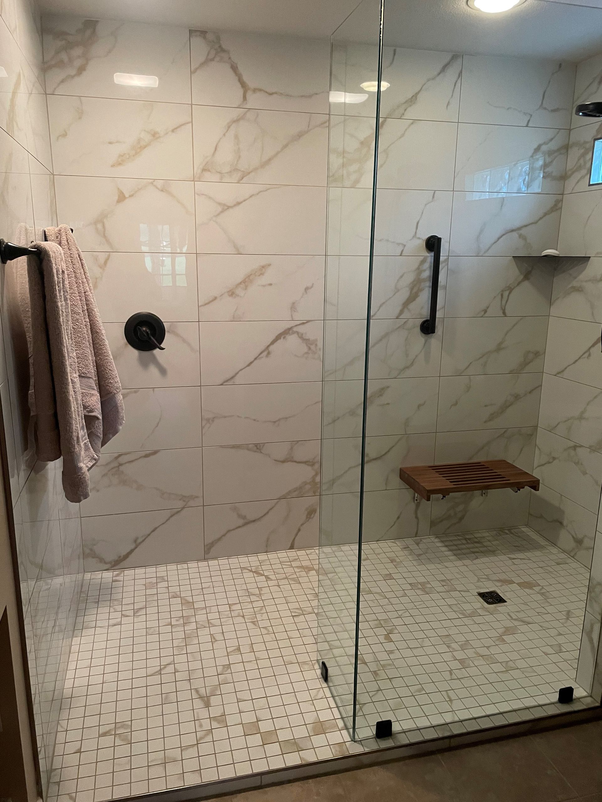 A modern walk-in shower with white and gold marble-patterned walls, small floor tiles, a glass door, and a wooden bench.