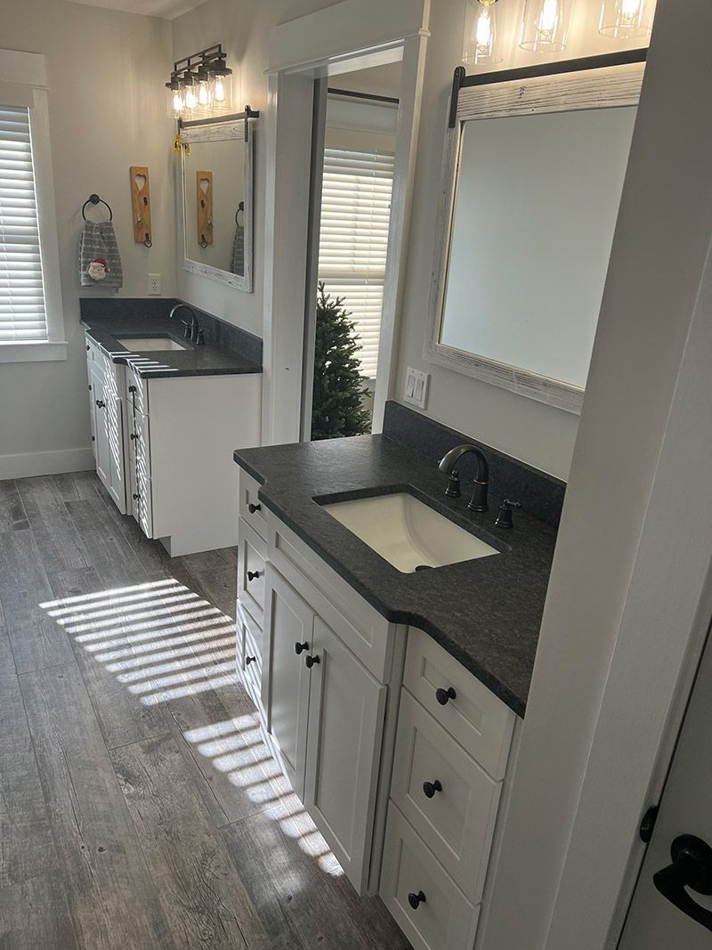 A bright, modern bathroom featuring white cabinets, dark gray countertops, dual sinks, mirrors, and wood-look flooring.