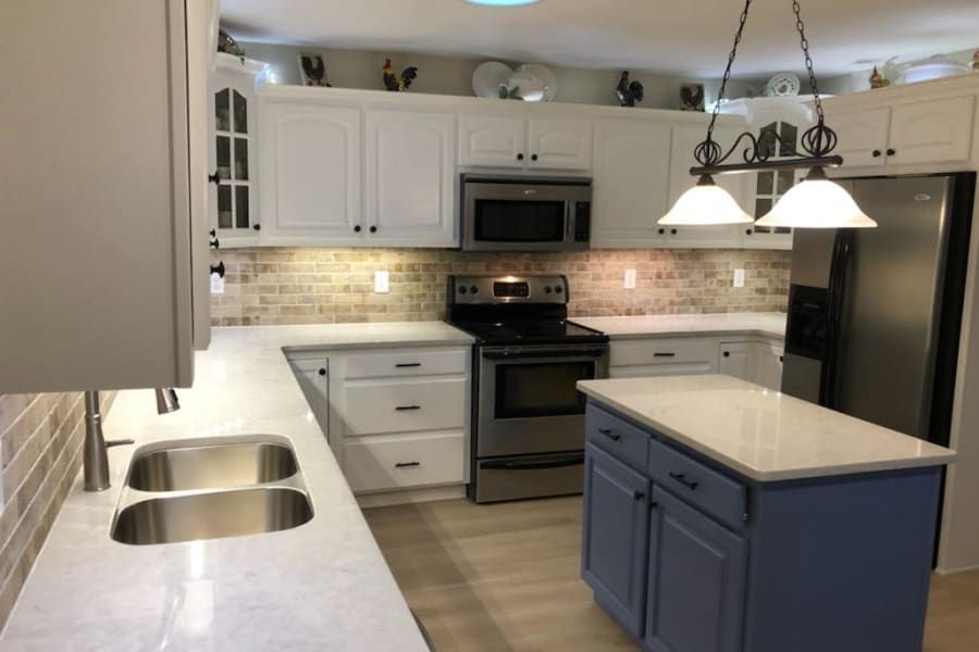 A kitchen with white cabinets, light countertops, stainless steel appliances, and a blue island under a hanging light.