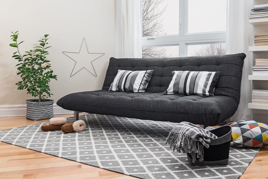 A dark grey futon on a geometric patterned rug in a bright room with a plant and a star wall decoration.