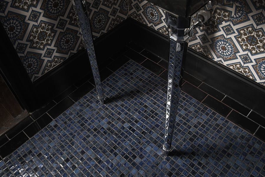 The silver legs of a sink stand on a dark blue mosaic-tiled floor against patterned wallpaper and black baseboards.