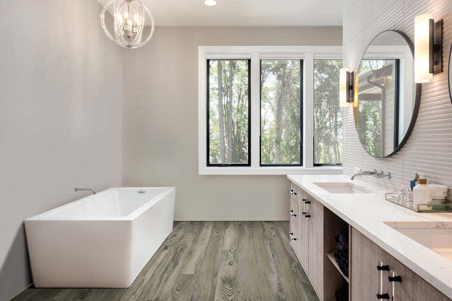 Modern bathroom with a white freestanding tub, wood-look flooring, a double vanity with a mirror, and large windows.