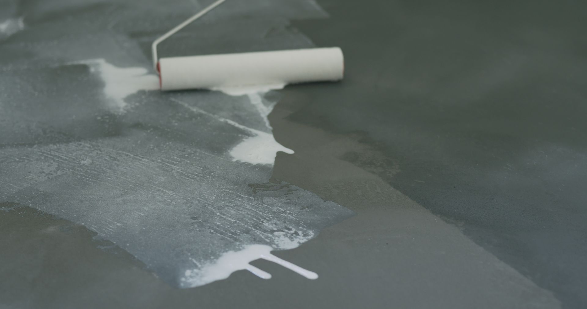 Paint roller applying white paint on a dark floor surface.