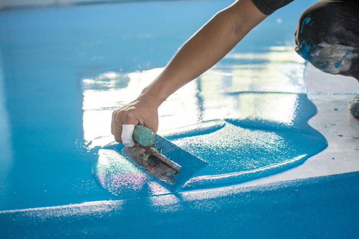 Hand smoothing bright blue paint over a glossy floor with a trowel