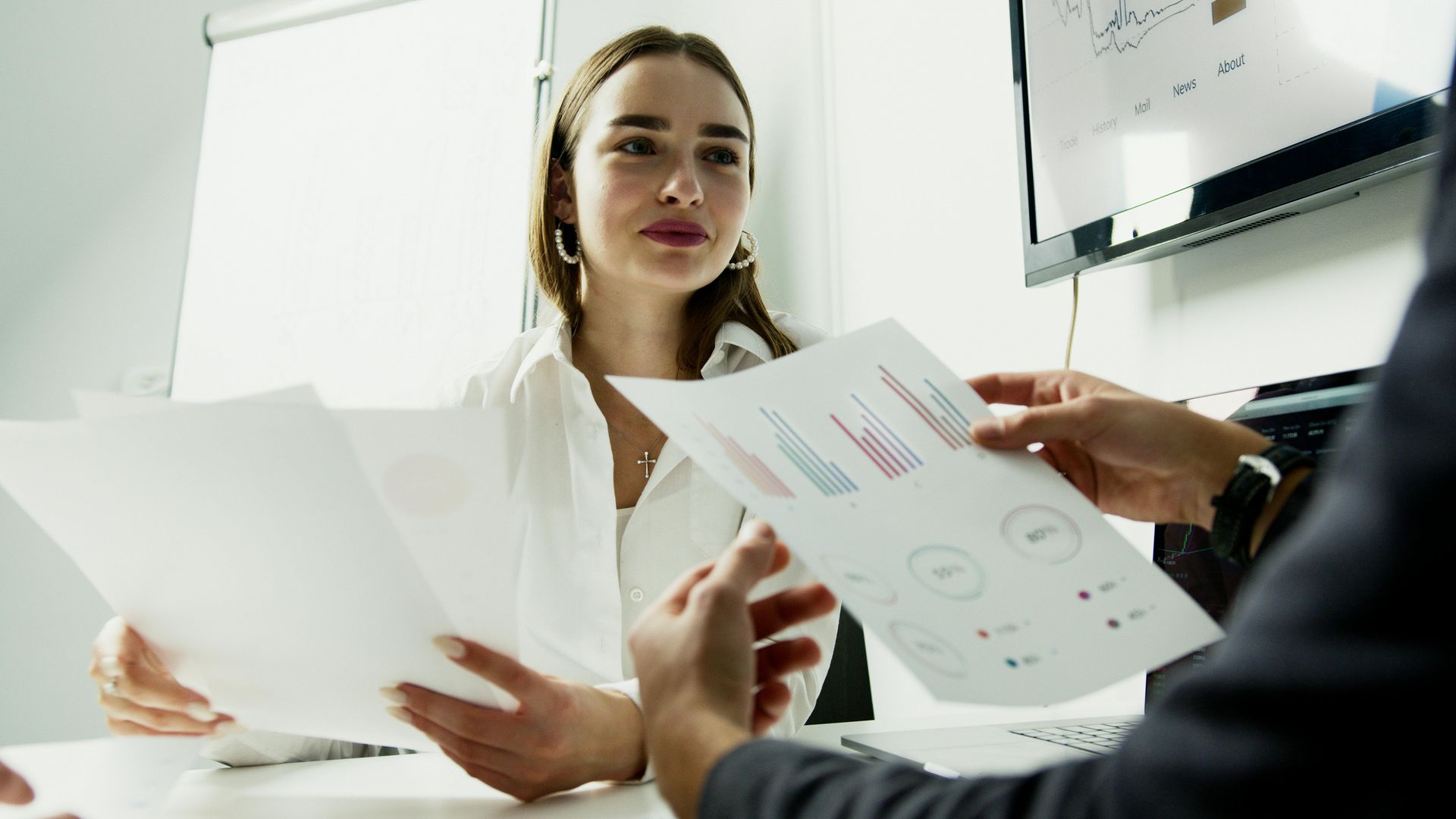 Woman reviewing papers with another person in a meeting setting; graphs and charts are visible.