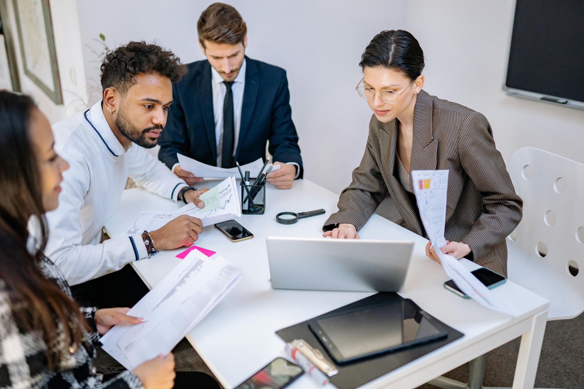 Four people in a meeting at a white table reviewing documents and a laptop, in an office.