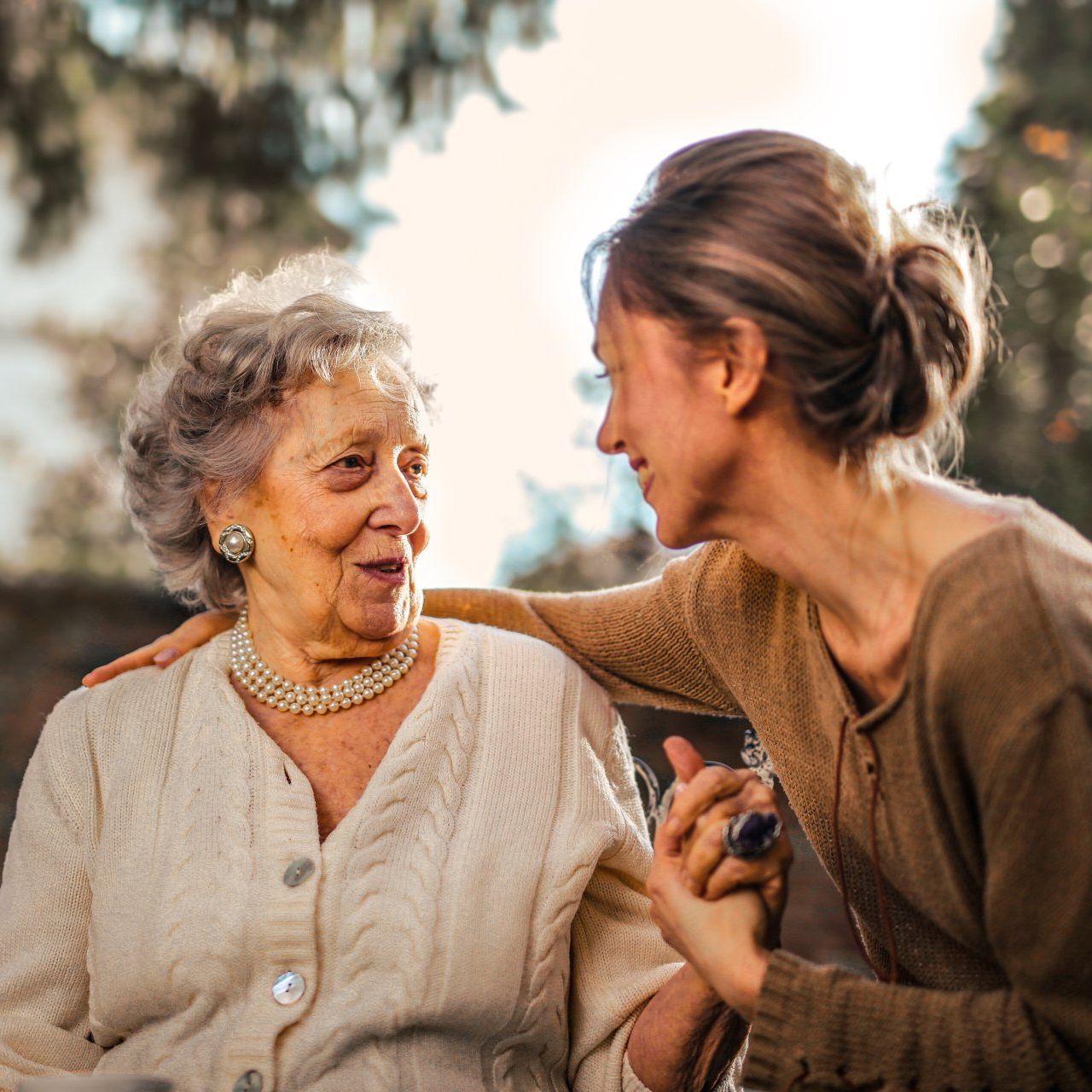 A person wearing a brown sweater holds the hands of a person in a white cardigan while they look at each other warmly.