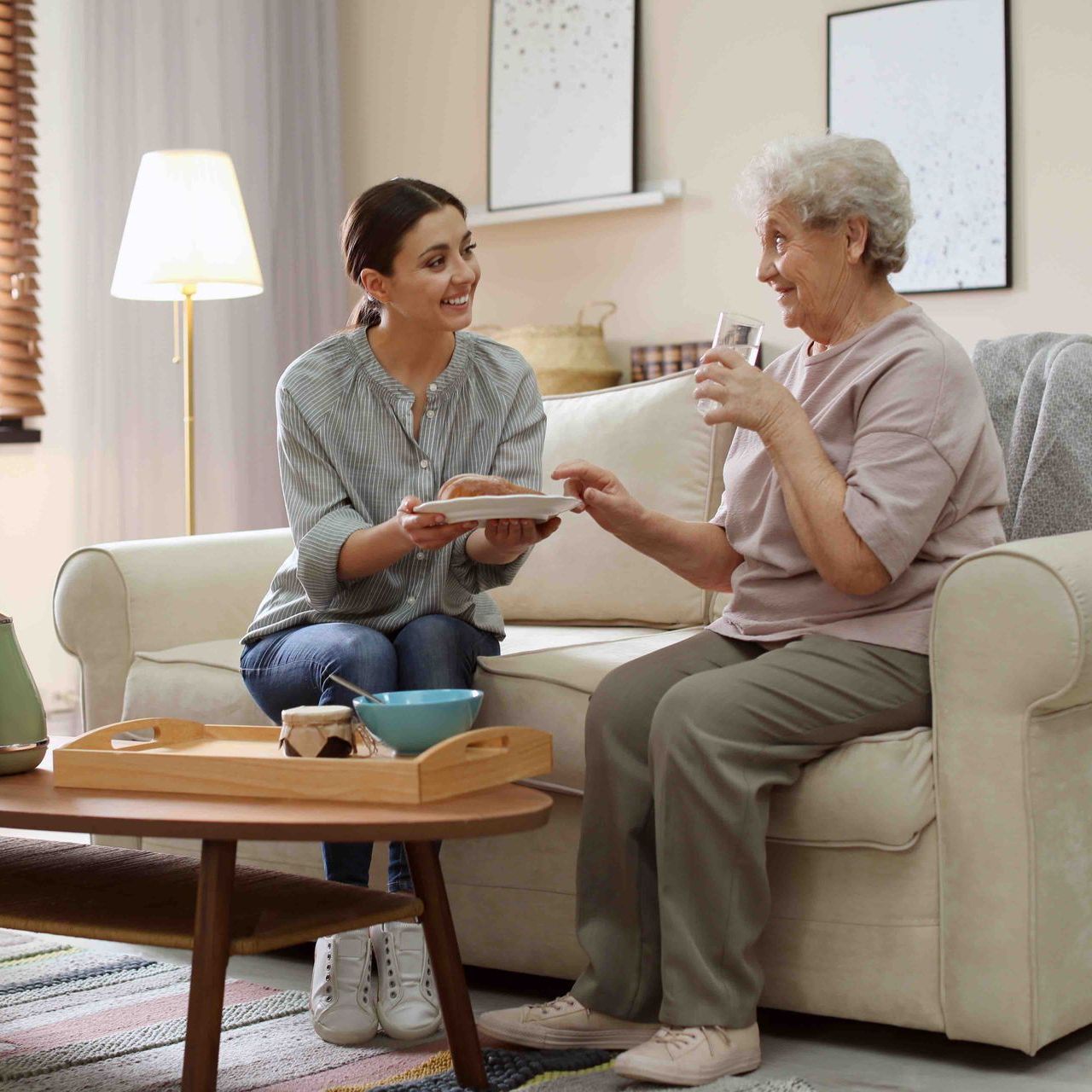 A caregiver offers a small plate to a person sitting on a sofa in a living room, while the person holds a glass of water.