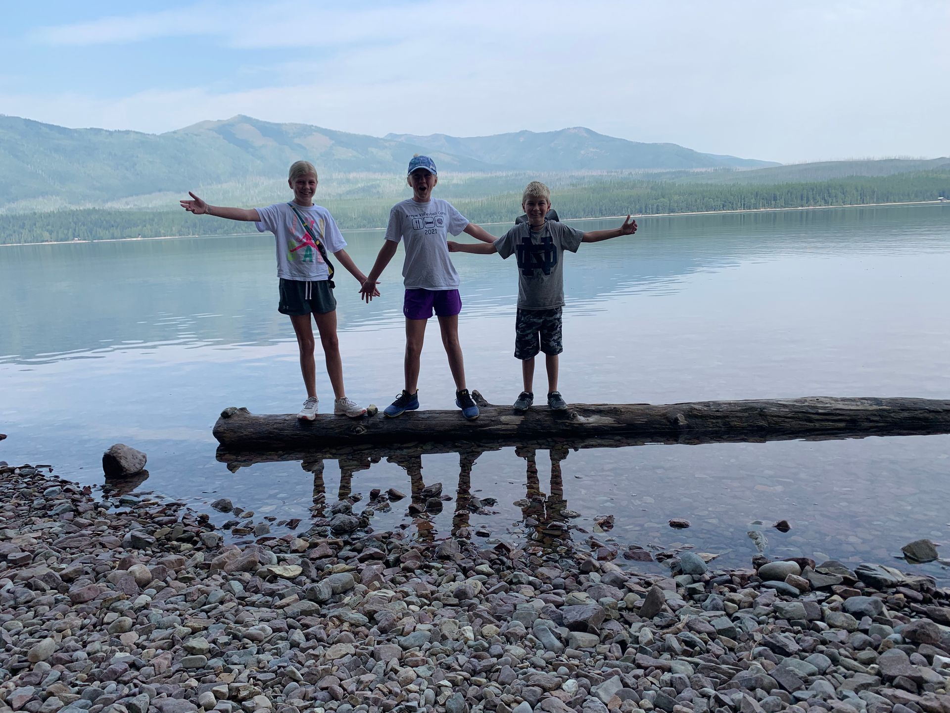 Three children are standing on a log by a lake holding hands.