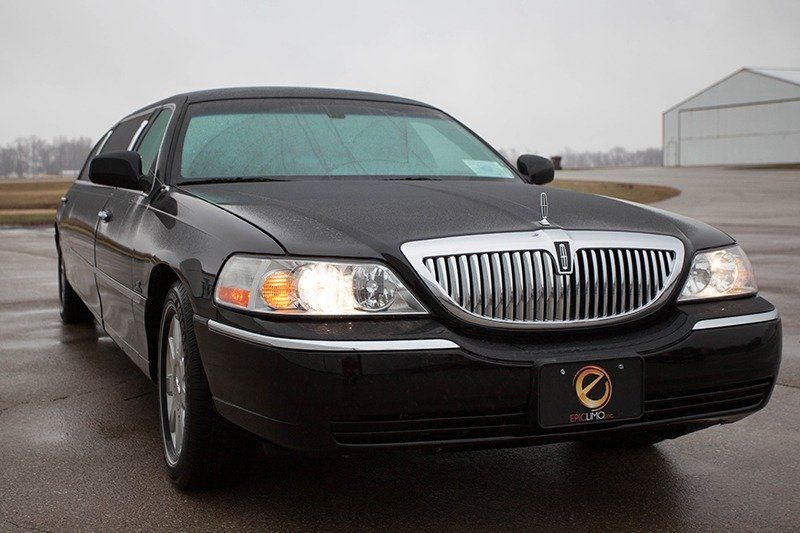 Black Lincoln Town Car limousine parked on a paved surface, with a cloudy sky in the background.