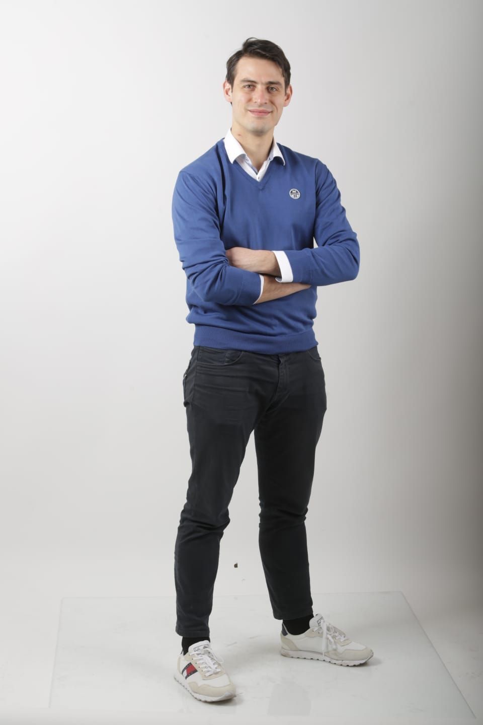 Un uomo con un maglione blu e pantaloni neri è in piedi con le braccia incrociate.