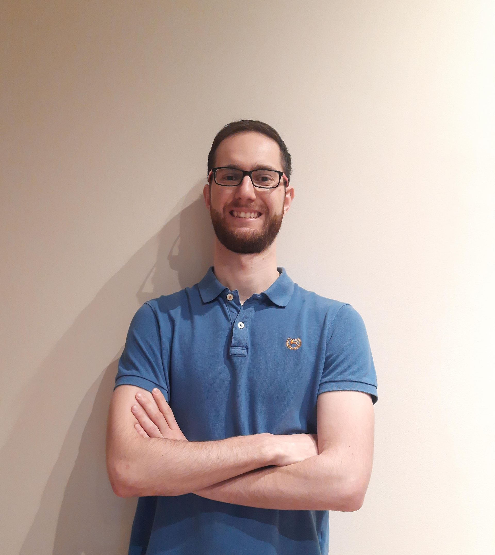 Un uomo con la barba, gli occhiali e una polo blu è in piedi con le braccia incrociate.