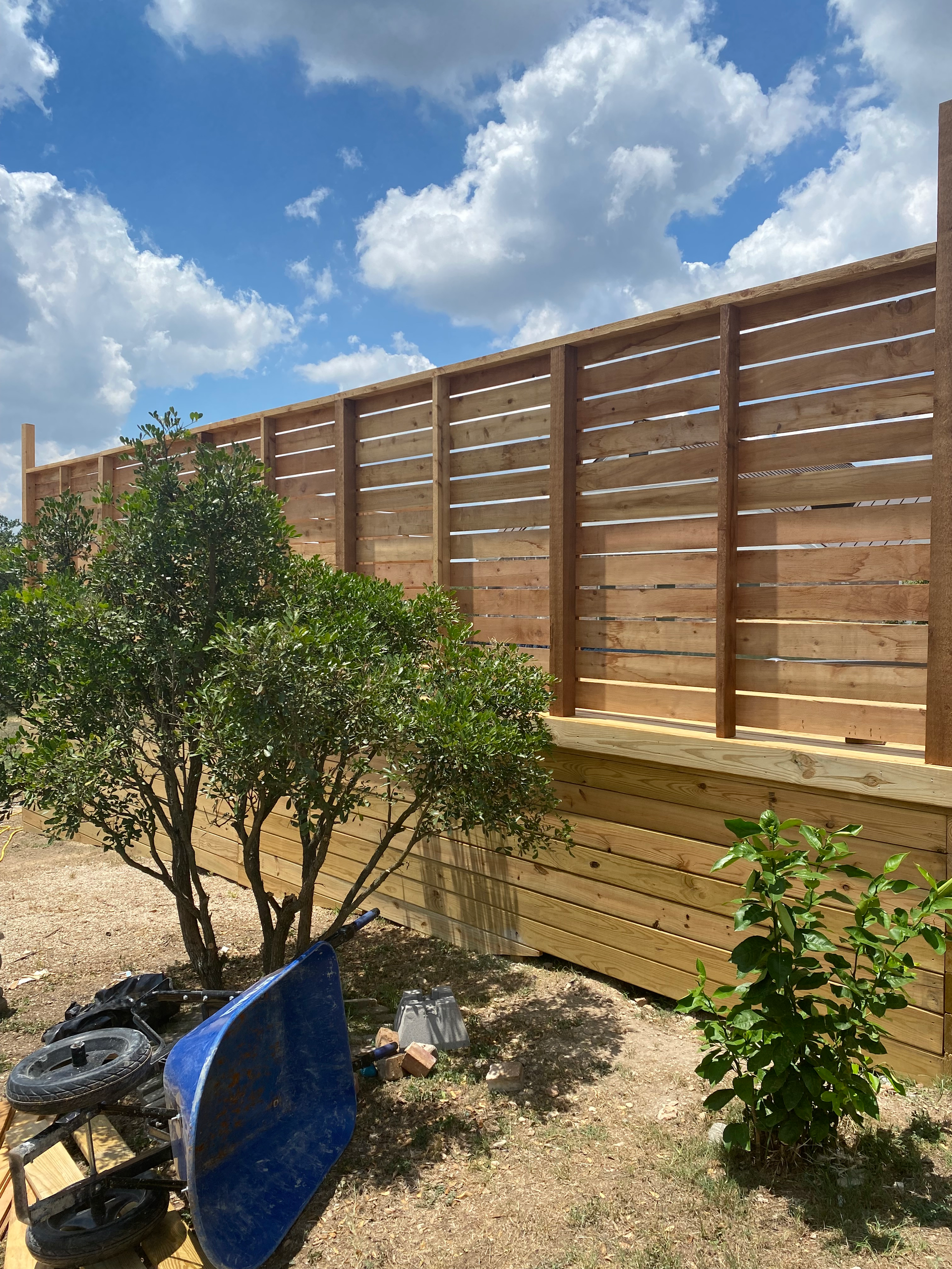 A wooden fence is surrounded by trees and a blue wheelbarrow.