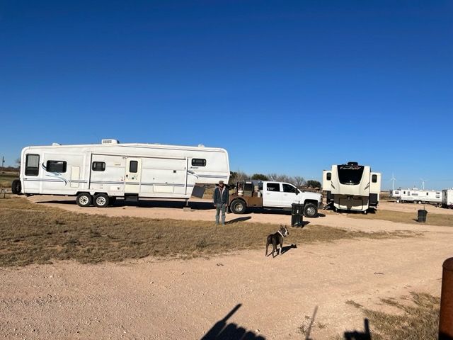 A man stands near a white truck towing a camper and another parked trailer. Dog in foreground on a sunny day.