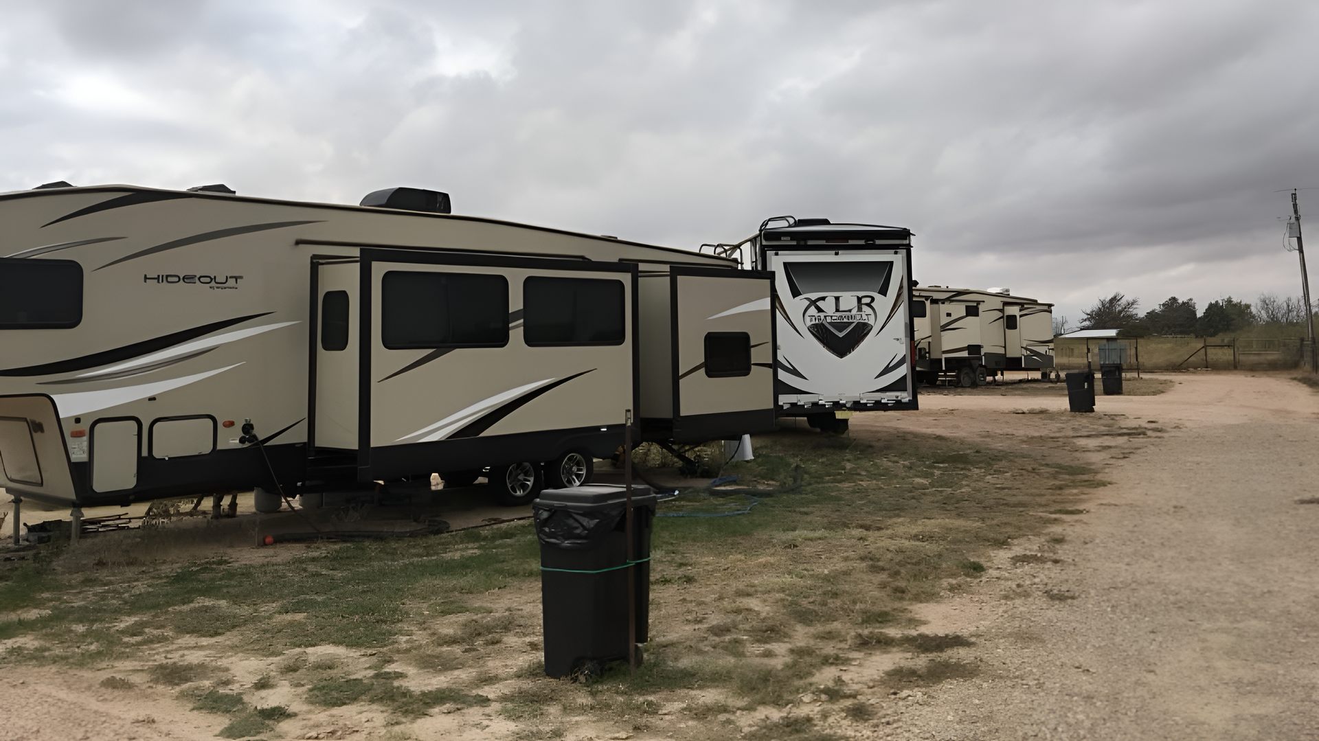 RVs parked at a campground on a cloudy day. Beige and white RVs line the gravel road, with trash cans nearby.
