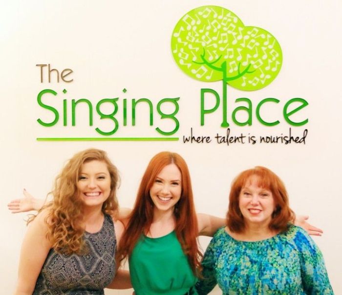 Three Woman Smiling Together in Front of the Singing Place Logo
