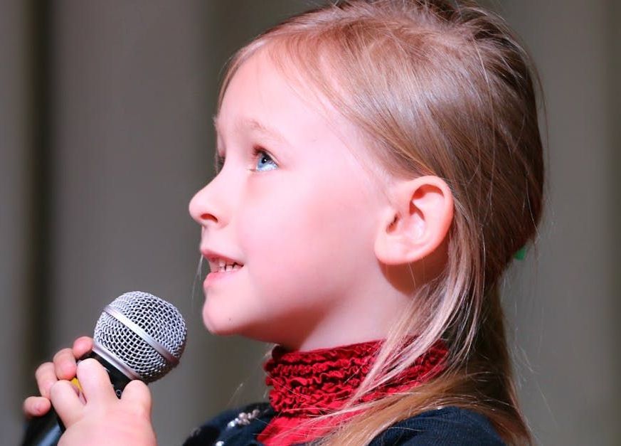 A little girl singing into a microphone in front of a wall that says the singing place children and teens singing in a choir