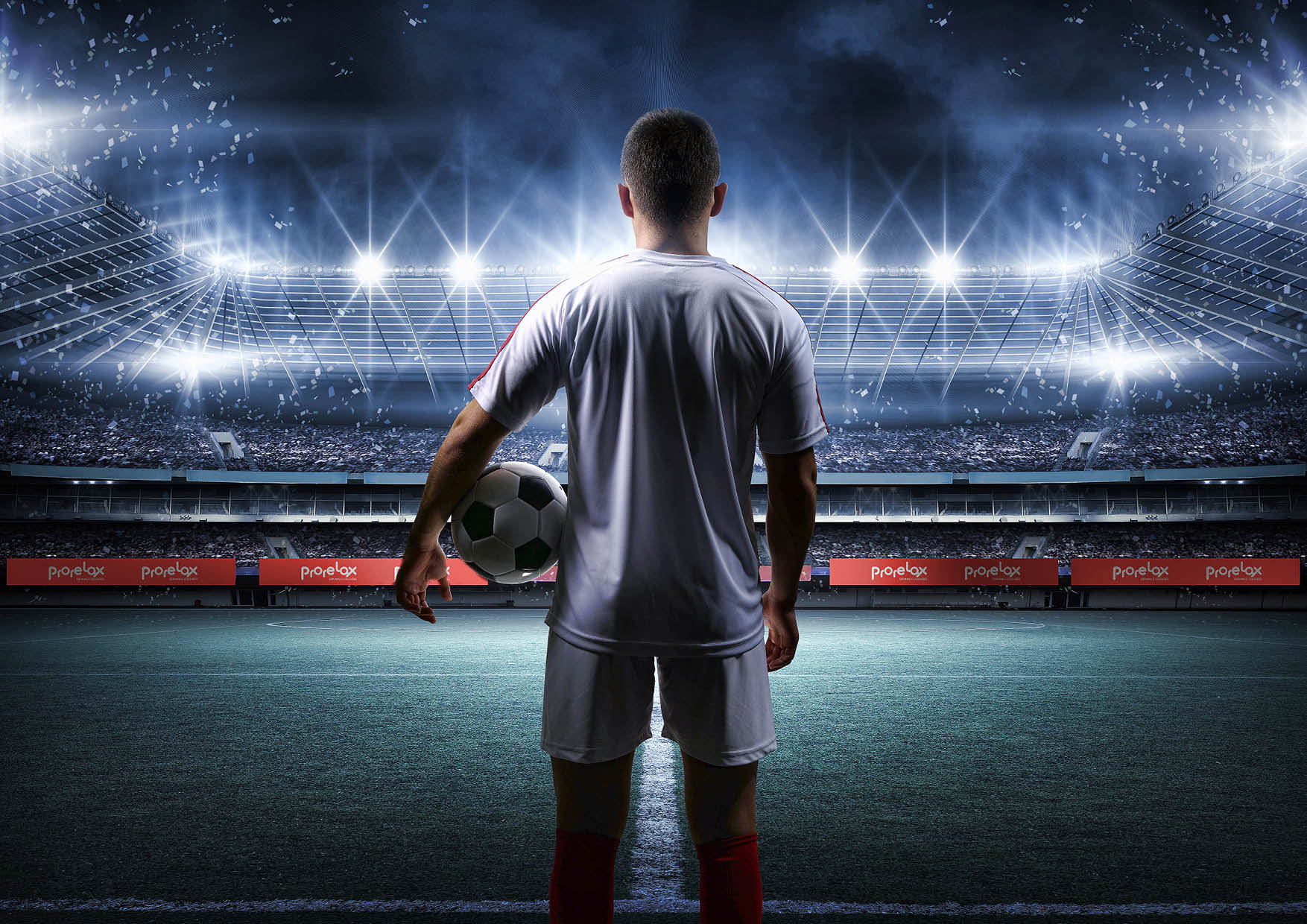 Jogador de futebol segurando uma bola, em pé no campo, de frente para um estádio lotado e iluminado por luzes brilhantes.