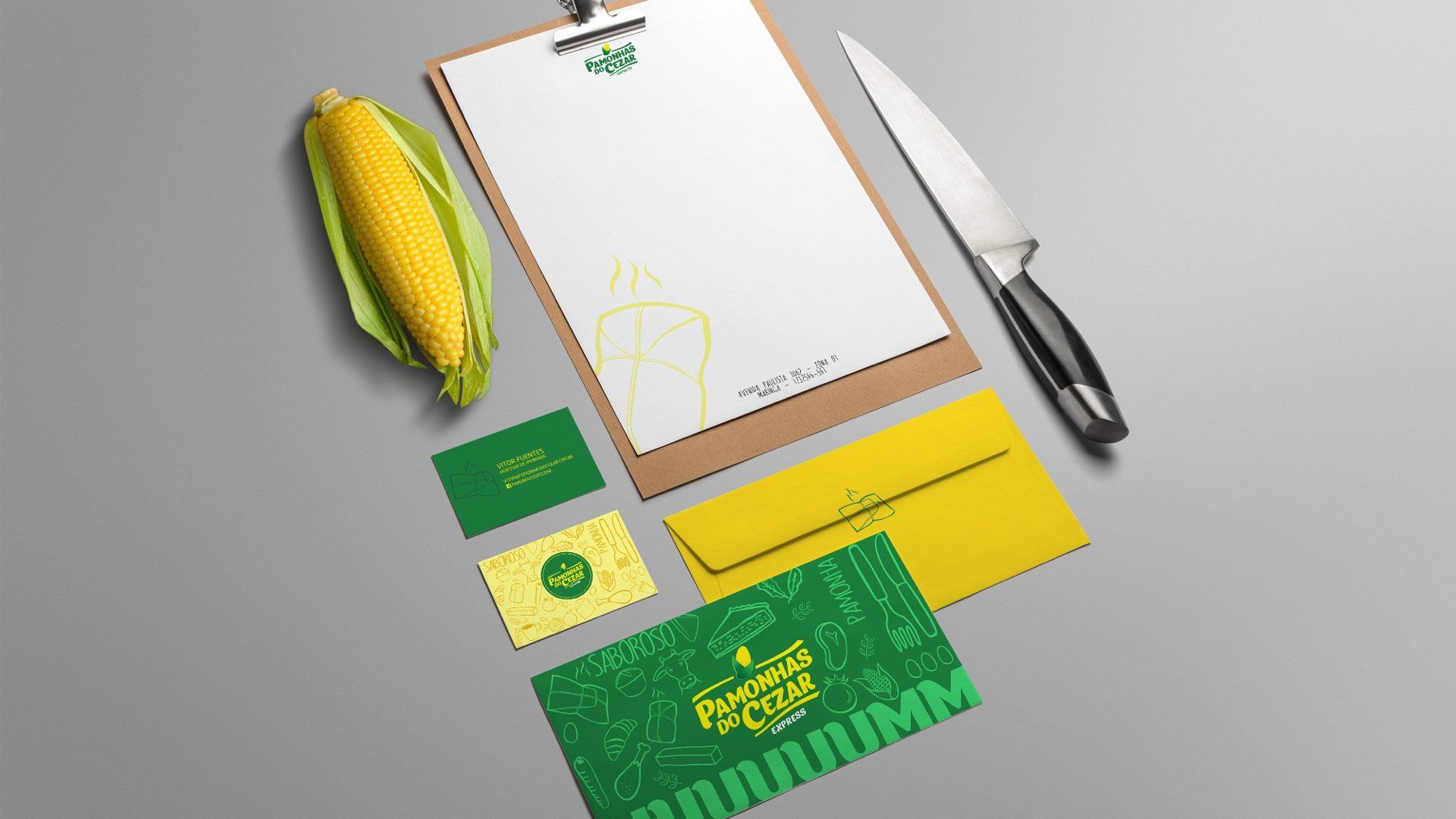 Maquete de papelaria com milho, uma faca e cartões de visita em tons de verde e amarelo sobre uma superfície cinza.