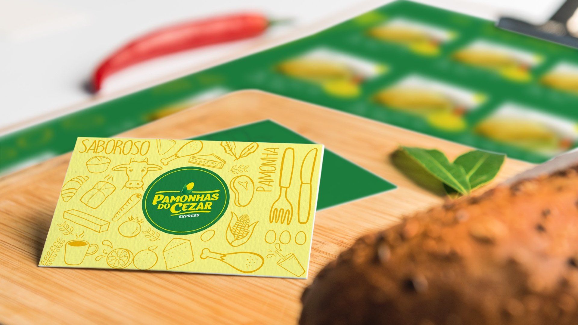 Cartão de visita sobre uma tábua de corte de madeira, com detalhes em verde e ilustrações relacionadas à culinária. Um pão desfocado e pimenta também aparecem na imagem.