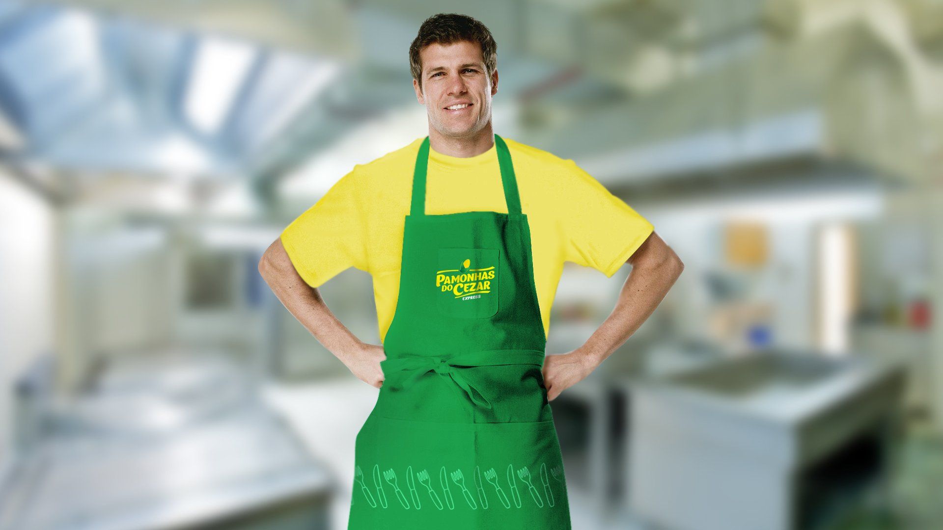 Um homem de camisa amarela e avental verde sorri, com as mãos na cintura, em uma cozinha comercial.