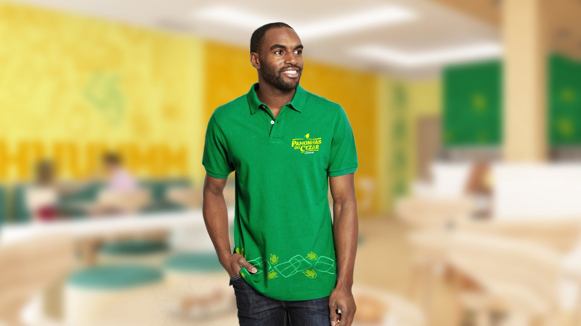 Um homem vestindo uma camisa polo verde com um logotipo sorri, em um ambiente interno. O fundo mostra um café amarelo e verde.