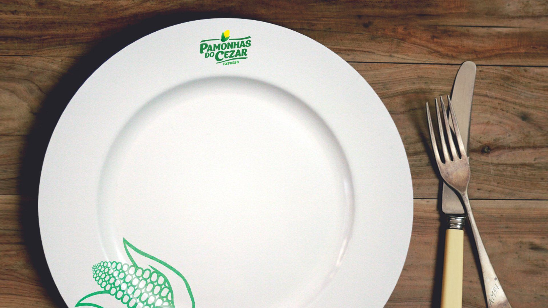 Prato branco com logotipo verde e ilustração de milho, acompanhado de faca e garfo sobre uma mesa de madeira.