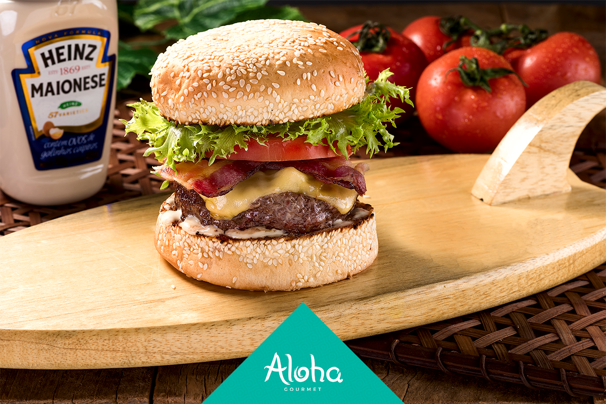 Hambúrguer em uma bandeja de madeira em formato de prancha de surfe com alface, tomate, bacon e queijo, com uma garrafa de maionese e tomates ao fundo.