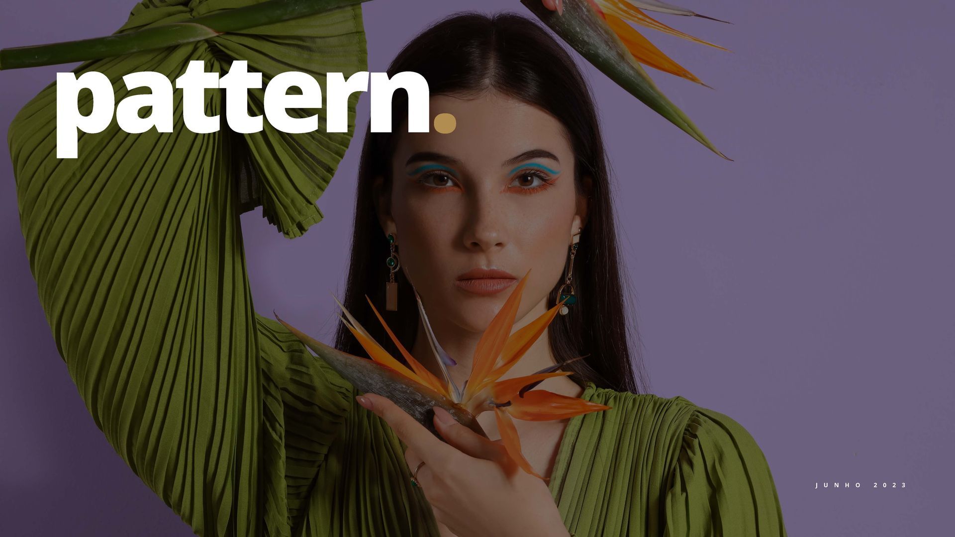 Mulher com longos cabelos escuros e maquiagem colorida nos olhos segura uma flor Ave do Paraíso em frente a um fundo roxo.