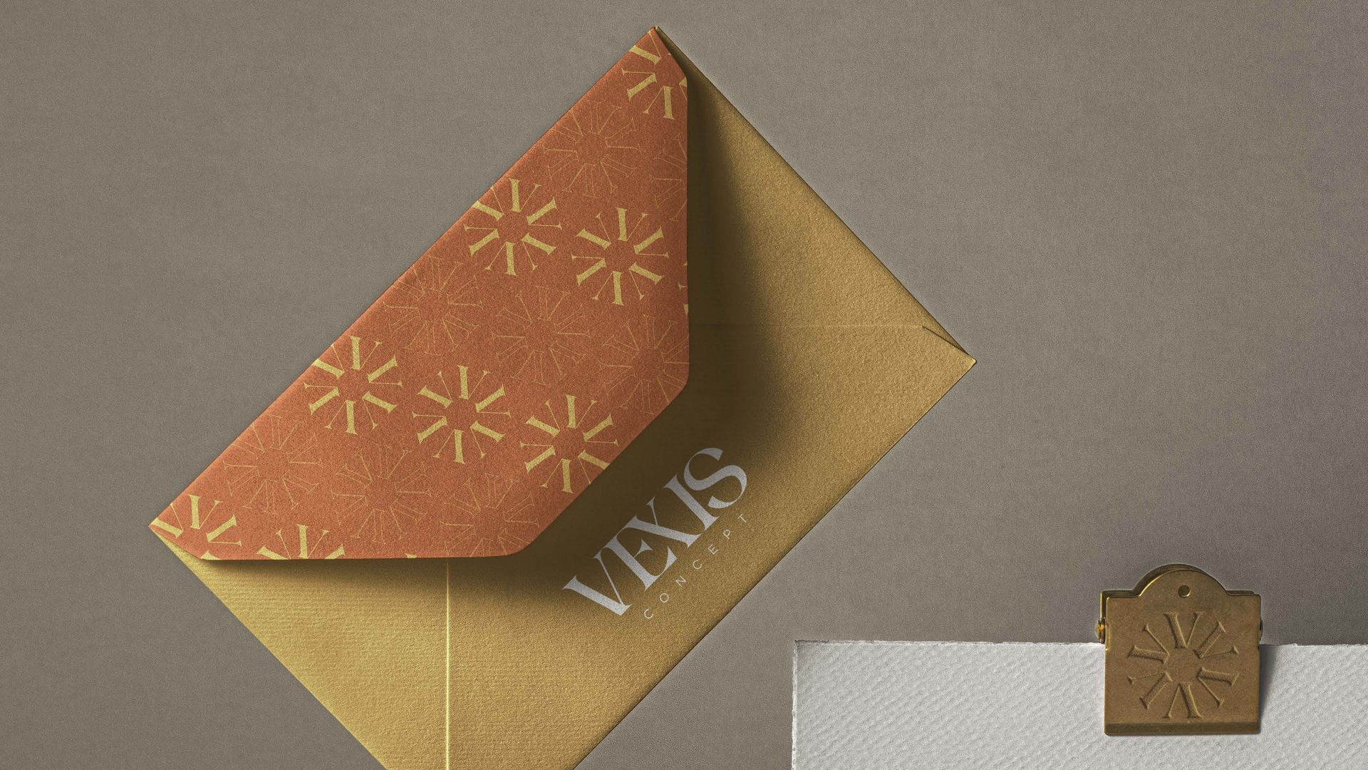 Um envelope dourado com uma aba estampada em laranja e um fecho decorativo sobre um fundo cinza.