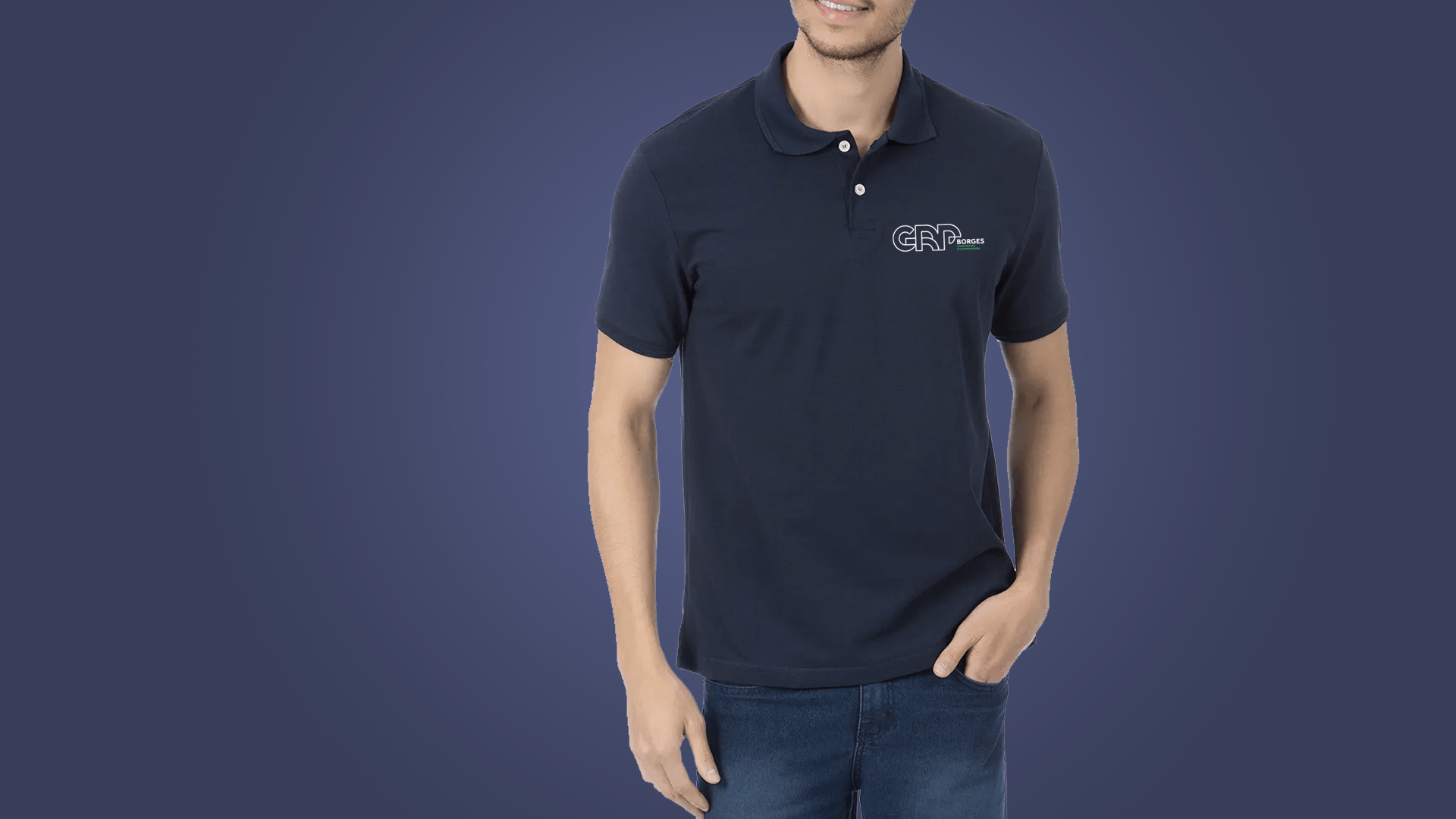 Homem vestindo camisa polo azul-escuro e calça jeans sorri, em pé, contra um fundo azul-escuro em degradê. A camisa tem um pequeno logotipo branco.