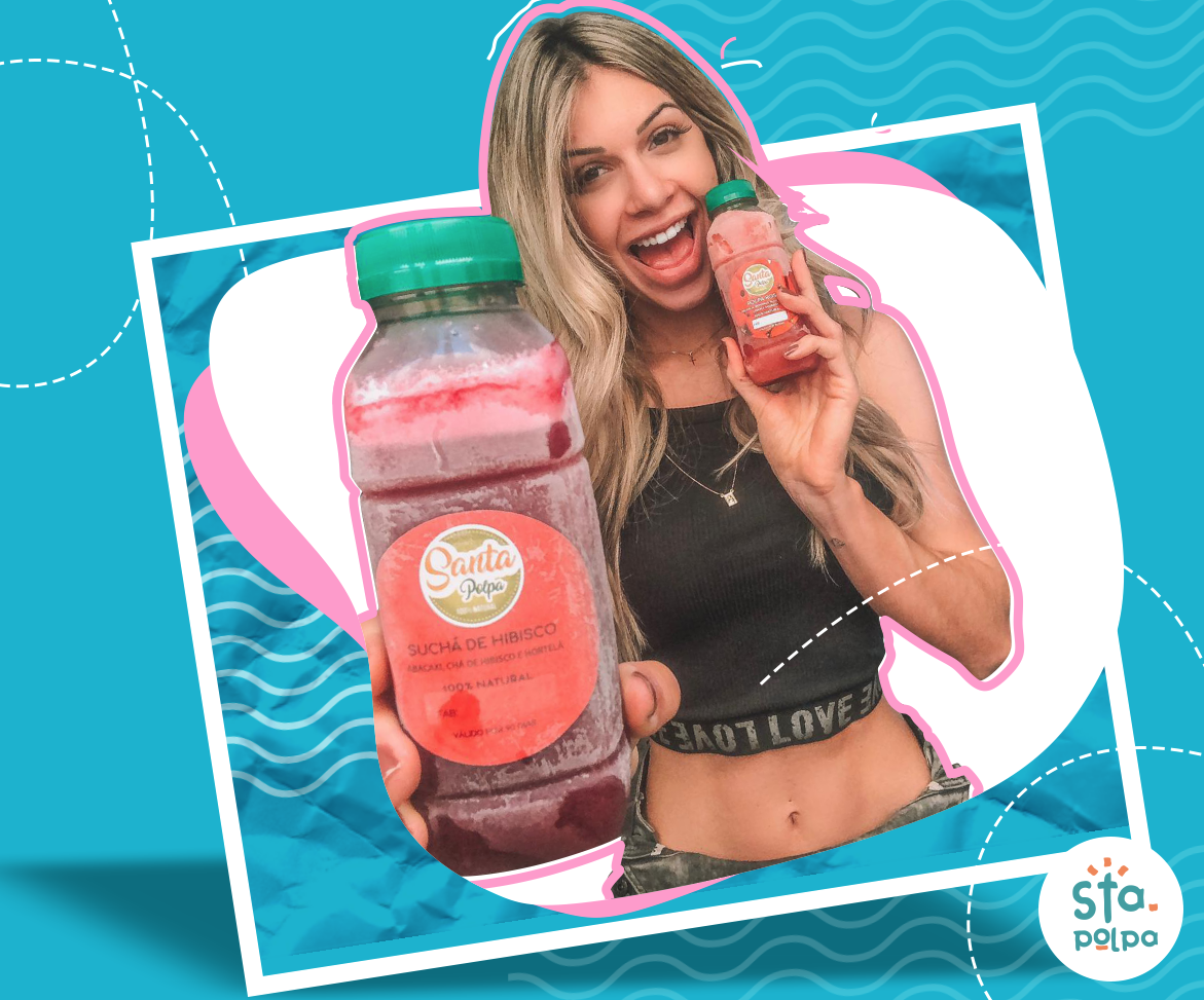 Mulher de cabelos loiros sorri, segurando uma garrafa grande e uma pequena de suco vermelho contra um fundo azul.