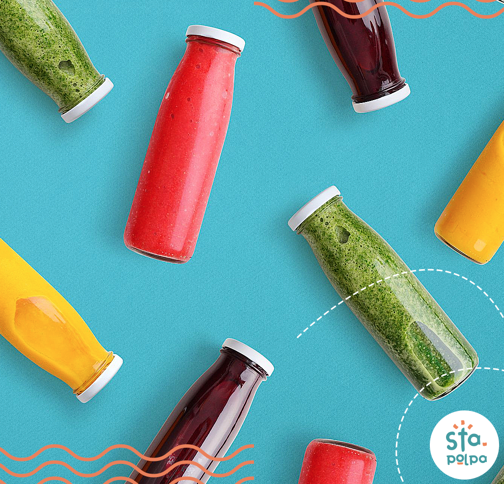 Garrafas de suco colorido dispostas em uma superfície azul, incluindo verde, vermelho, amarelo e roxo.