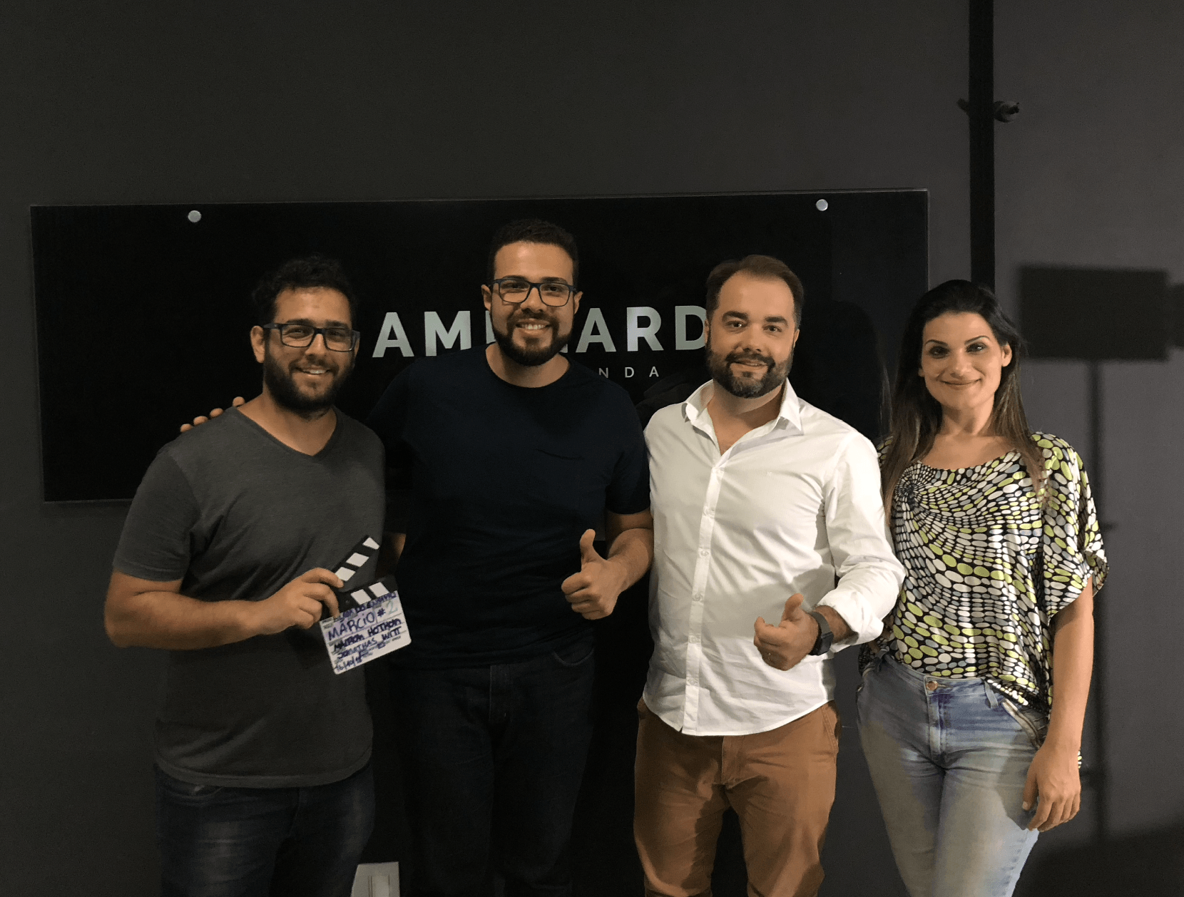 Quatro pessoas posam em frente a uma placa preta. Elas sorriem e seguram uma claquete.