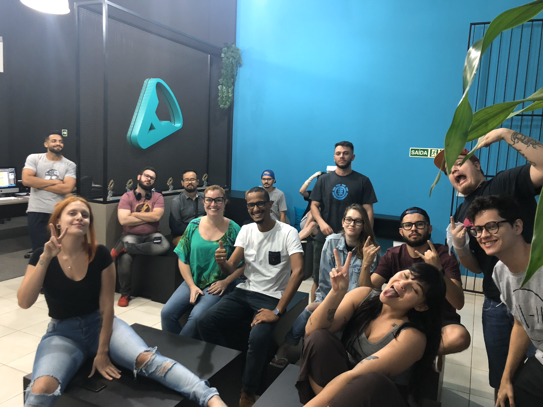 Grupo de pessoas diversas em um escritório colorido; algumas estão sorrindo, fazendo sinais de paz e posando para a câmera.